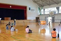 体育館でオレンジ色のボールカゴの周りを大きく取り囲むようにして床に座っている児童の写真