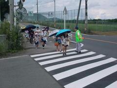 屋外で傘をさして横断歩道を渡ろうとしている複数の児童たちと、それを見つめている一人の大人の写真