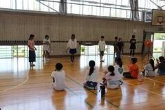 体育館で数人の児童が立っていて、そちらを座っている数人の児童が見つめている写真