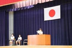 舞台の上の壇上で1名の子どもが紙を見てを読み上げており、横には椅子に座った子どもが2名いる写真