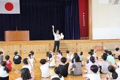体育館の舞台の前で大人が右手を挙げながら左手に白いものを持ち、それを床に座っている子どもたちが眺めている写真