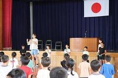 体育館の舞台の前に幾人かの子どもが立っており、そちらの方を向いた大勢の子どもたちが立っている写真
