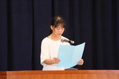 室内の演壇でマイクを使い、見開きの用紙を持ち、話をしている児童の写真