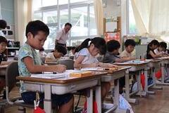 学校の教室で子供たちが整列した机にの椅子に座り勉強をている写真