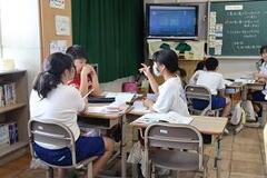 学校の教室で3人の子供たちが机をくっつけて手ぶりを交えて会話をしている写真