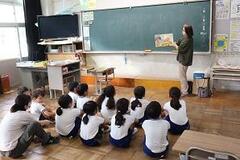 学校の教室で子供たちが体育座りをしている前で女性が本を持って中身を見せている写真