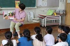 学校の教室で子供たちが体育座りをしている前で女性が中身を開いた絵本を手に持って見せている写真