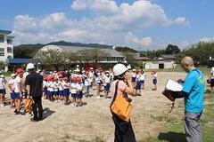 校庭で大人たちがヘルメットを被ったりファイルを見ている中で体操服に紅白帽姿の子供たちが整列している写真