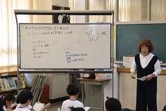 室内の文字が書かれたホワイトボードの隣で女性が話をしているのを子どもたちが座りながら聞いている写真