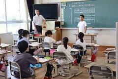学校の教室で男の子が黒板の前に立ち、席に座った児童たちがそれを見ている写真
