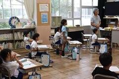 学校の教室で中央を空けて並べた机にの椅子に座った子どもたちの内、一人が立っている写真