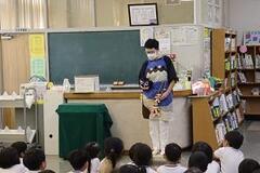 室内で床に座った子どもたちの前で男性がエプロンをして立っている写真