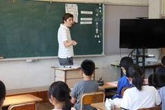 学校の教室で黒板の前で男性が話をしており、席に座った子どもたちがそれを見ている写真