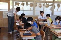 学校の教室で子供たちが席に座っており、男性が1人の席の前で様子を見ている写真