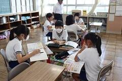 学校の教室で3人の子供たちが机を合わせて本を見ながら話合っている写真