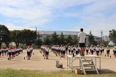 学校の校庭で子供たちが体操服姿で整列しており、それと向かい合った朝礼台に男性が立っている写真