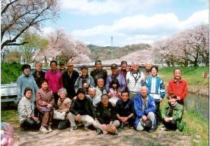 桜咲く川辺で記念撮影する東沢1区の皆さんの写真