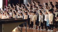 体育館のステージ前に沢山の児童たちが並び、片手を上に挙げている写真