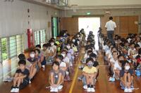 体育館のような所に沢山の児童が体育座りをしながら並んでいる写真
