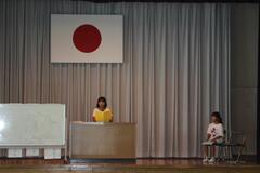 日本国旗のようなものがぶら下がったステージ上に、講演台があり1人の児童が立っており、その右隣に1人の児童が椅子に座っている写真