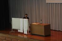 ステージ上に講演台とホワイトボードのようなものがあり、ホワイトボードの後ろに1人の男性が立っている写真