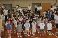 体育館のような所に、沢山の児童達と複数の大人が立っている写真