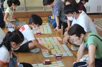 床にカルタのようなカードが幾つか並べられており、児童と大人が向かい合わせて座っている写真