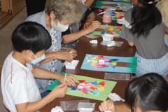 児童と女性が画用紙の上に折り紙を貼って作品を作っている写真