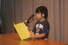 演台で1人の児童が原稿のようなものを手に持ち、マイクに向かって話をしている写真