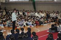 体育館のような所で沢山の児童が座っている中で1人の児童がマイクを持って立っている写真