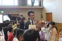 児童達が両端に立っている間を花束を持った男性が通っている写真