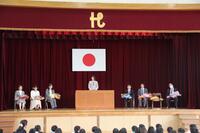 体育館壇上で退任予定の先生が1人ずつ挨拶をしている様子の写真
