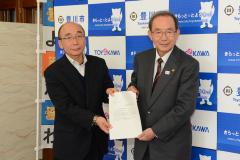 豊川市のロゴやキャラクターが書かれた背景の前にスーツの男性2人が一緒に紙を持って並んでいる写真