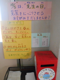 赤い箱型のにっこりポストとミニはがきが置かれた机と、先生の日とお手紙の説明が書かれた張り紙の写真