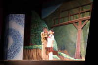 劇団の公演での、木と山のセットを背景にした和装の人物と女子学生の写真