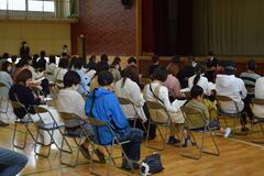 体育館に並んだパイプ椅子に座る大人たちの写真