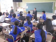 教壇の前で立っている人とそれを眺めている児童たちの写真