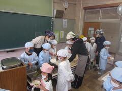 エプロンと三角巾を着けた大人たちと一緒に、給食着を着て給食を配膳する子供たちの写真