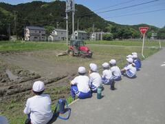 稲の植わっていない田んぼに置かれたトラクターを田んぼわきから眺める子供たちの写真