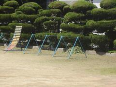 丸く刈り込まれた植木の前に逆上がりの補助台が付いた鉄棒の置かれた広場の写真