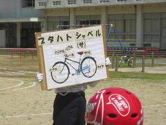 自転車の絵の描かれた看板を持っている人を見ている赤いヘルメットを着けた人の写真