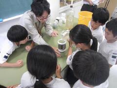 机に置かれた装置の説明をする大人とその周りに集まる子供たちの写真