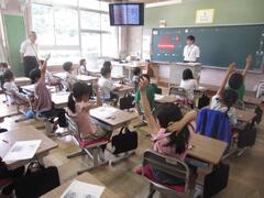 2人の大人のいる教室で手を挙げている複数の子供たちの写真