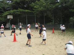 バスケットゴールと三角コーンの置かれた広場で体操着姿の子供たちが歩き回っている写真