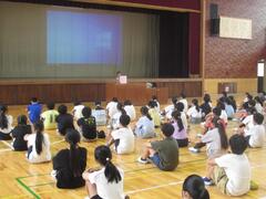 体育館の床に並んで座り、前に立つ大人の話を聞く子供たちの写真