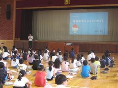 体育館のステージ奥の幕に映し出された資料を床に座って見ている子供たちの写真