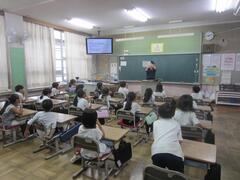テレビの点いた教室で話す大人を見ている子供たちの写真