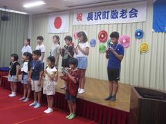 「祝 長沢町敬老会」と書かれた横断幕の飾られたステージでリコーダーを吹く子供たちの写真