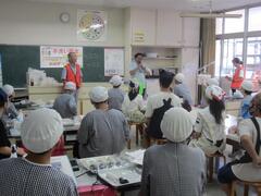 給食帽子をつけた子供たちが大人の話を聞いている写真