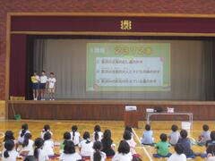 体育館のステージの端に立つ3人と幕に映った資料を見る複数の子供たちの写真